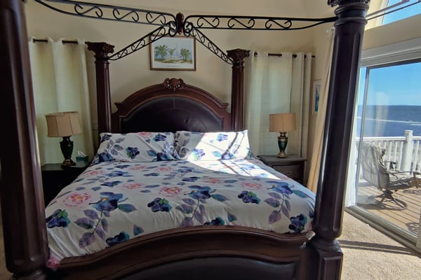 Master bedroom with dark wood four-poster bed, floral bedding, ocean view through glass doors, and oceanfront deck with railing.
