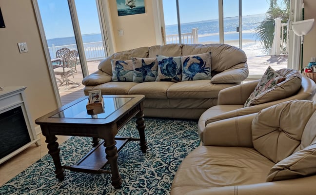 Beachfront living room with tan leather sofa and armchair, ocean view through glass doors, nautical-themed pillows, and wooden coffee table.