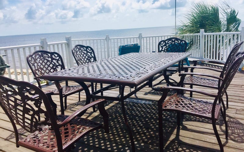 Oceanfront deck with metal dining table, ornate chairs, white railing, and palm tree overlooking calm sea under blue sky