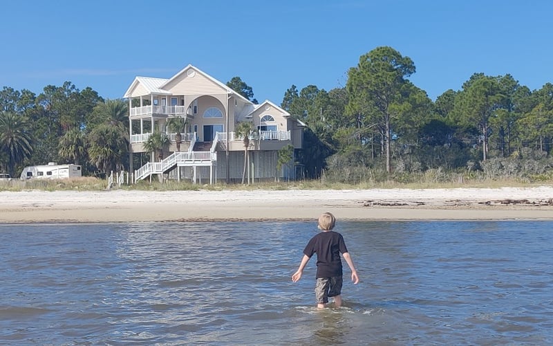 Child wading in ocean water with elevated beachfront house on stilts behind sandy shore and pine trees