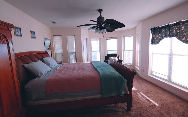 Spacious bedroom with red upholstered bed, ceiling fan, multiple windows with ocean view, and pink walls.
