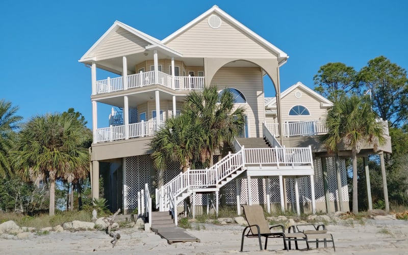 Elevated white beach house with multiple decks and palm trees, lounge chairs on sandy beach below